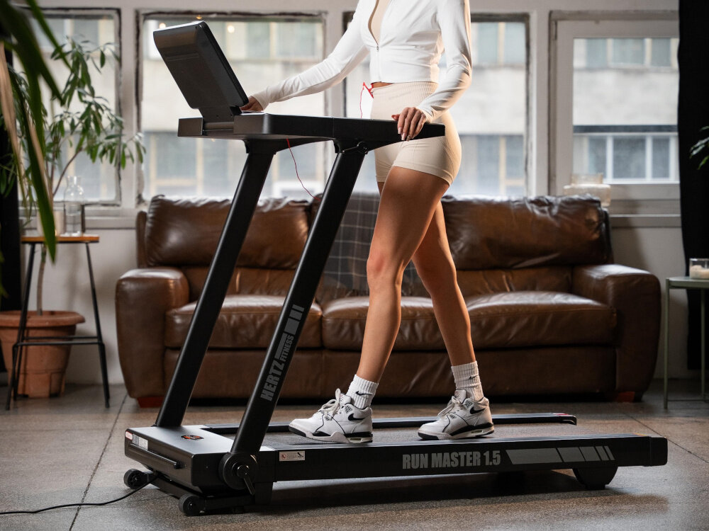 Osoba ćwiczy na bieżni elektrycznej HERTZ FITNESS Run Master 1.5 w jasnym pomieszczeniu z dużymi oknami. Na bieżni widoczne są napisy \'HERTZ FITNESS\' i \'RUN MASTER 1.5\'.