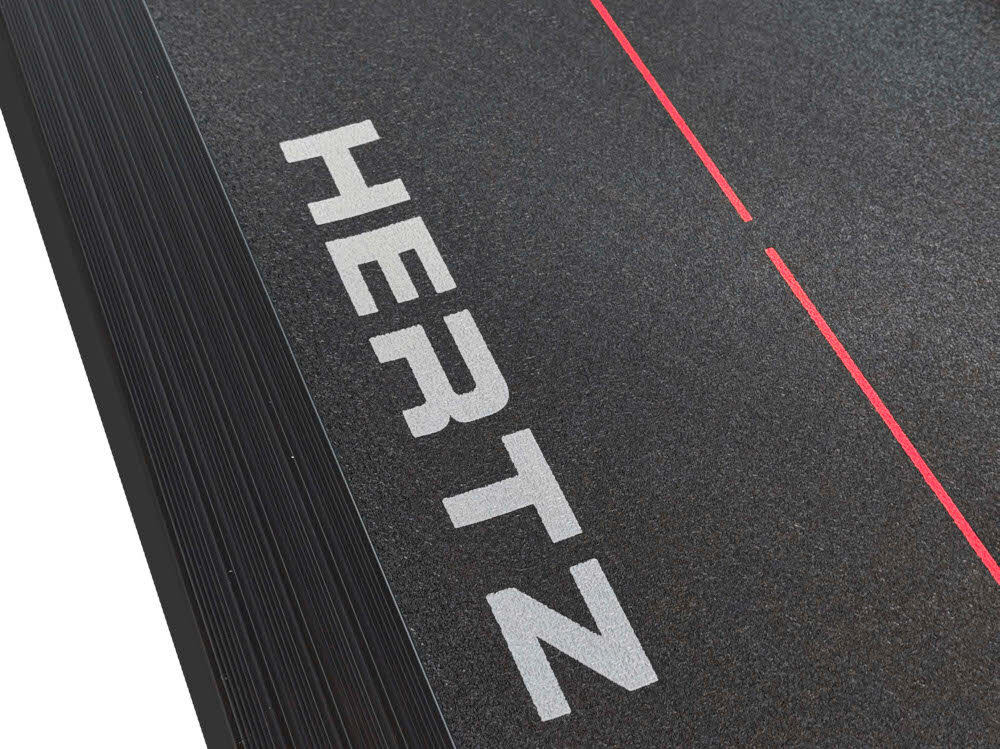 Fragment bieżni z widocznym napisem \'HERTZ\' na ciemnej powierzchni oraz czerwonymi liniami dekoracyjnymi.