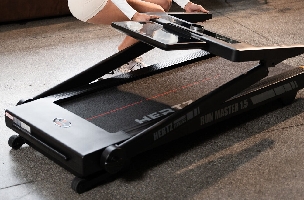Na obrazie widoczna jest bieżnia elektryczna z napisem \'HERTZ FITNESS RUN MASTER 1.5\'. Osoba w jasnym stroju sportowym ustawia panel sterujący bieżni.