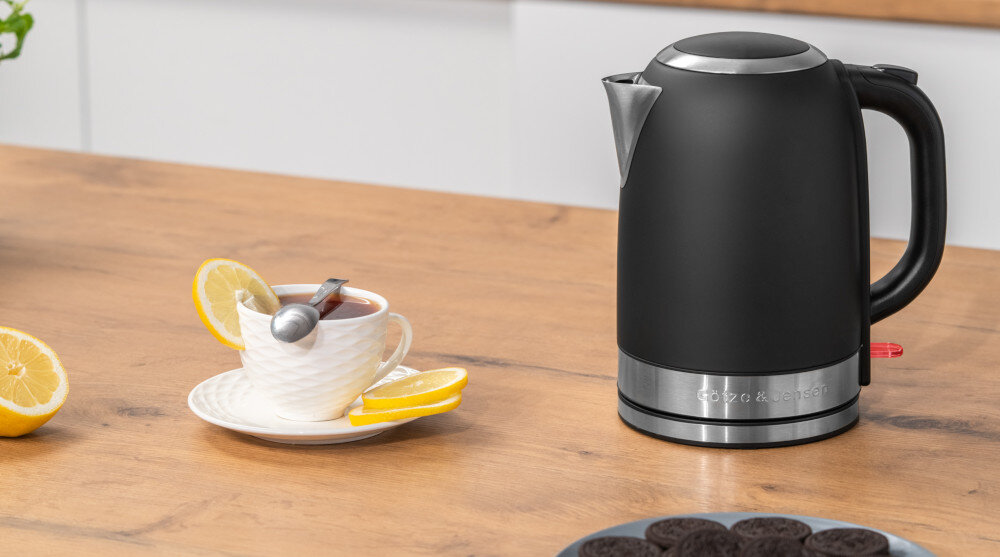 Na drewnianym blacie stoi czarny czajnik elektryczny z metalowym wykończeniem i napisem \'GÖTZE & JENSEN\'. Obok znajduje się filiżanka herbaty z plasterkami cytryny oraz talerz z ciastkami.