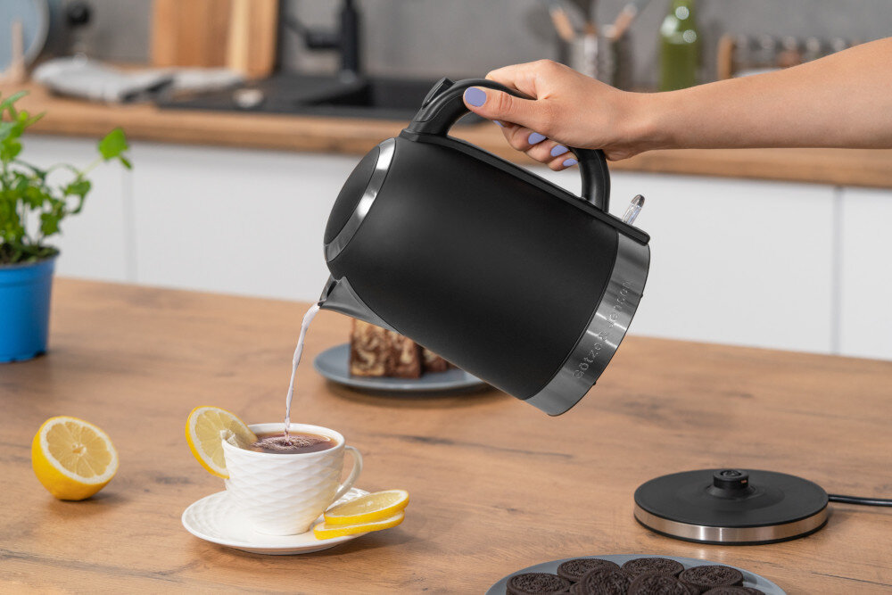 Osoba nalewa wodę z czarnego czajnika do filiżanki z herbatą, obok której leży plasterek cytryny. Na stole znajdują się również ciastka i pokrywka czajnika.