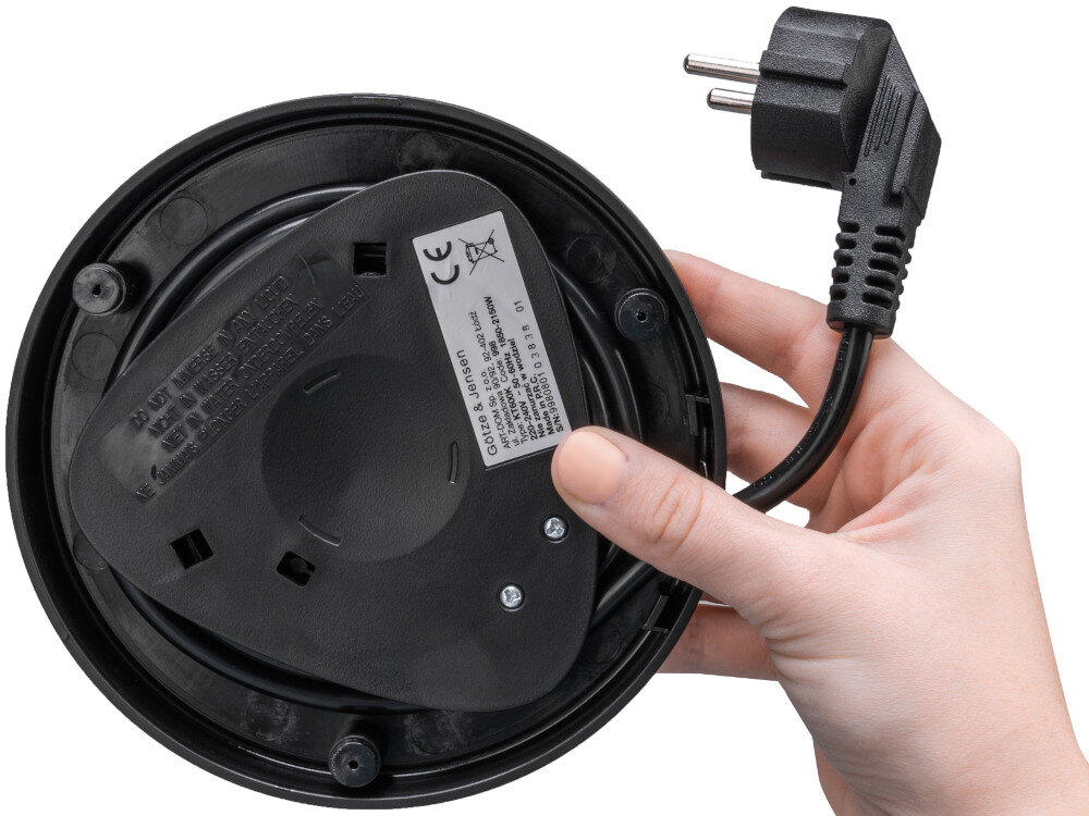 Osoba trzyma podstawę czajnika elektrycznego z widocznym przewodem zasilającym i wtyczką. Na podstawie znajduje się etykieta z napisem \'Götze & Jensen\'.