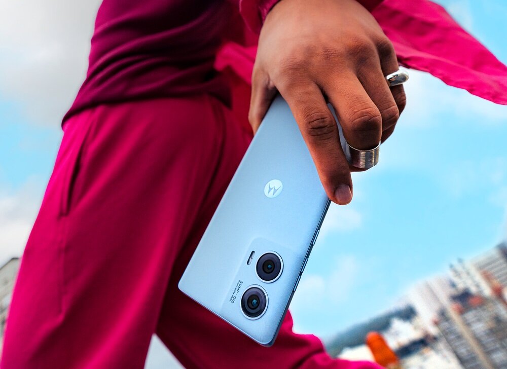 Osoba trzyma smartfon z logo Motorola, w kolorze niebieskim, z dwoma widocznymi obiektywami aparatu. W tle widać fragmenty budynków i niebo.