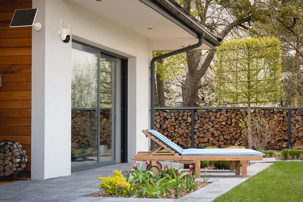 Na tarasie przy domu znajduje się drewniany leżak z niebieskim materacem. W tle widać stos drewna oraz roślinność, a na ścianie zamontowana jest kamera i panel słoneczny.