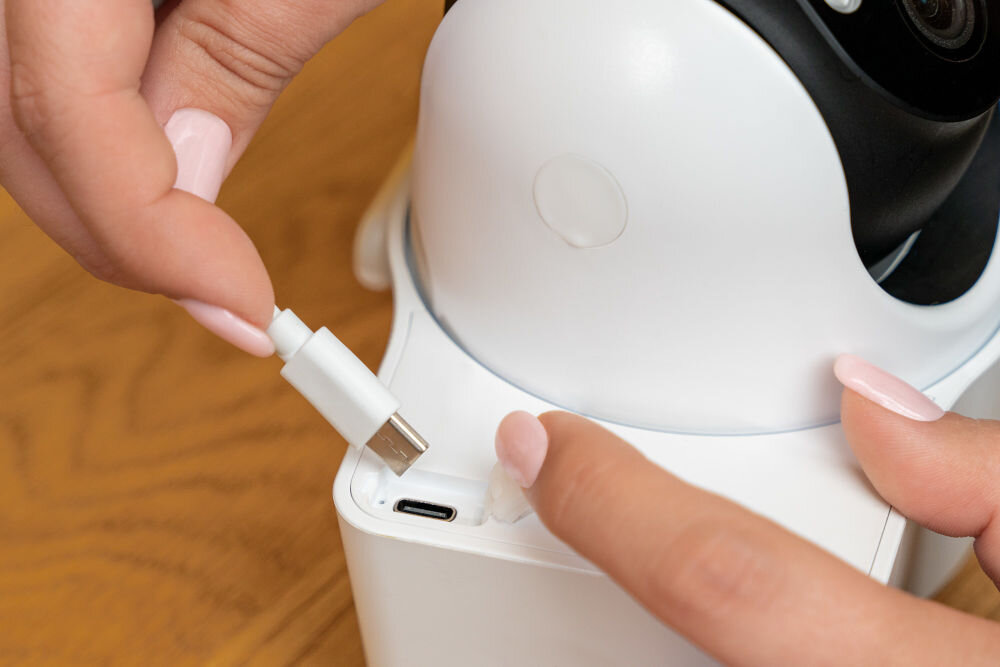 Osoba podłącza kabel USB do białego urządzenia, które wygląda na kamerę obrotową. Kamera ma czarny obiektyw i znajduje się na drewnianej powierzchni.