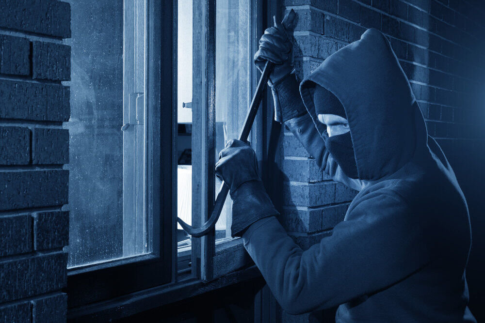 Osoba w kapturze i masce próbuje otworzyć okno za pomocą łomu. Scena jest oświetlona na niebiesko, co nadaje jej nocny charakter.