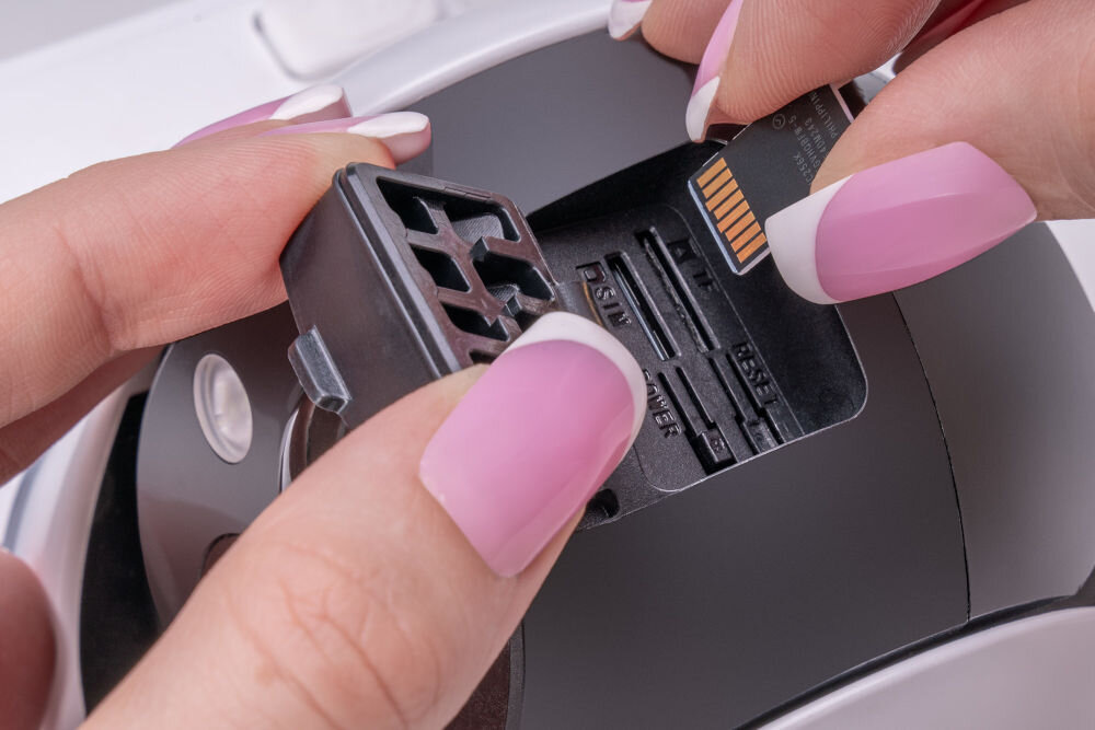 Osoba wkłada kartę microSD do gniazda w urządzeniu elektronicznym. Widoczne są dłonie z różowym manicure oraz część obudowy urządzenia.