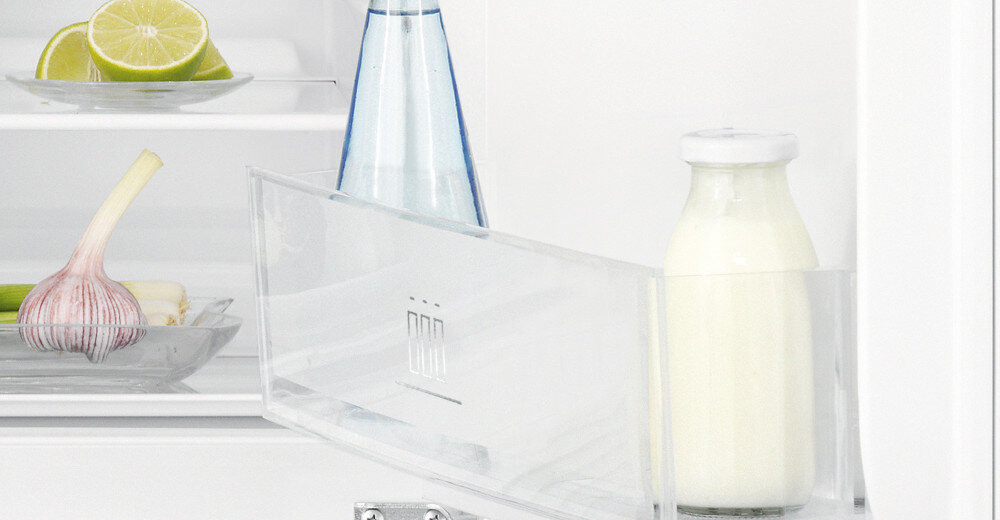 Wnętrze lodówki z widoczną półką, na której stoi butelka z mlekiem i przezroczysta butelka z niebieską etykietą. W tle na półce znajduje się talerz z pokrojoną limonką i główką czosnku.