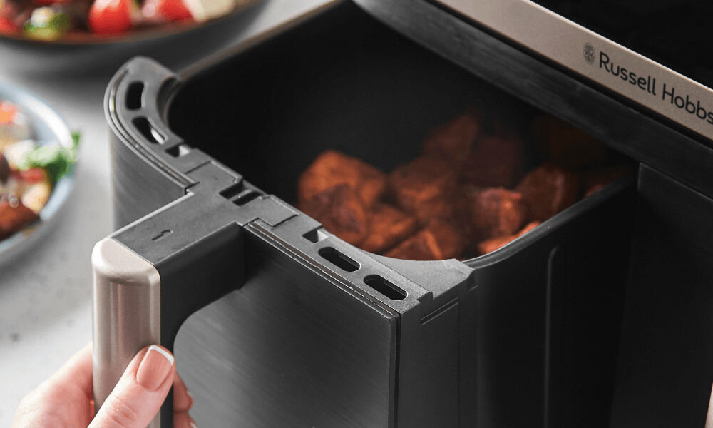 Osoba trzyma uchwyt szuflady frytkownicy beztłuszczowej, w której znajdują się kawałki jedzenia. Na urządzeniu widoczne jest logo \'Russell Hobbs\'.
