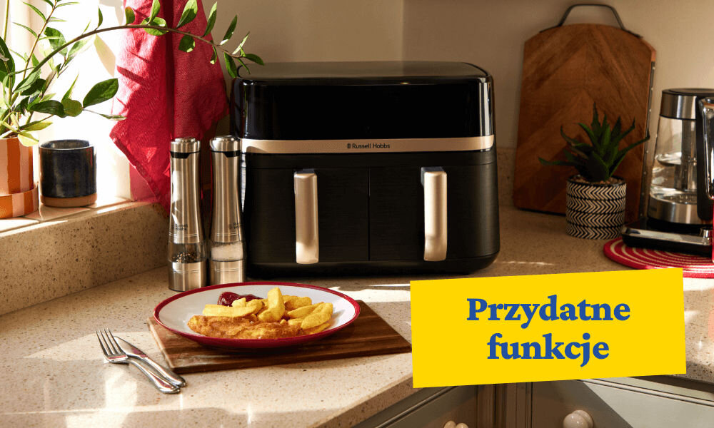 Na blacie kuchennym stoi czarna frytkownica beztłuszczowa z dwoma uchwytami i logo \'Russell Hobbs\'. Obok znajduje się talerz z frytkami i rybą, a na żółtym tle widnieje napis \'Przydatne funkcje\'.
