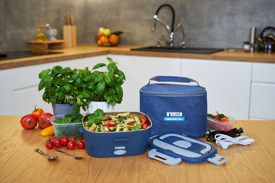 Na drewnianym blacie kuchennym znajduje się elektryczny lunch box NOVEEN z jedzeniem, obok torba termiczna z logo NOVEEN oraz świeże warzywa i zioła. W tle widoczny jest zlew i akcesoria kuchenne.
