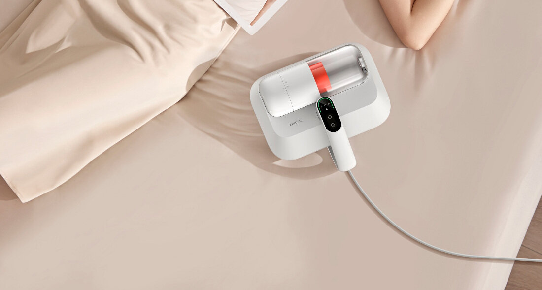 Na jasnym tle znajduje się biały odkurzacz z przezroczystym pojemnikiem i widocznym logo \'XIAOMI\'. Obok leży osoba, której widoczna jest część ciała.