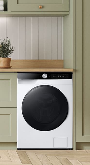 Pralko-suszarka w białym kolorze z czarnym panelem sterowania i dużymi drzwiczkami. Urządzenie stoi w kuchni z zielonymi szafkami i drewnianym blatem.