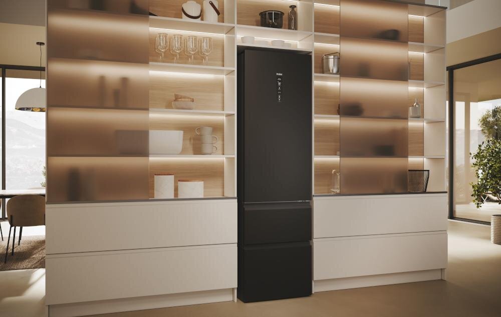 Czarna lodówka umieszczona jest pomiędzy nowoczesnymi półkami z oświetleniem, na których znajdują się szklanki i dekoracje. W tle widoczne są duże okna wpuszczające naturalne światło do pomieszczenia.