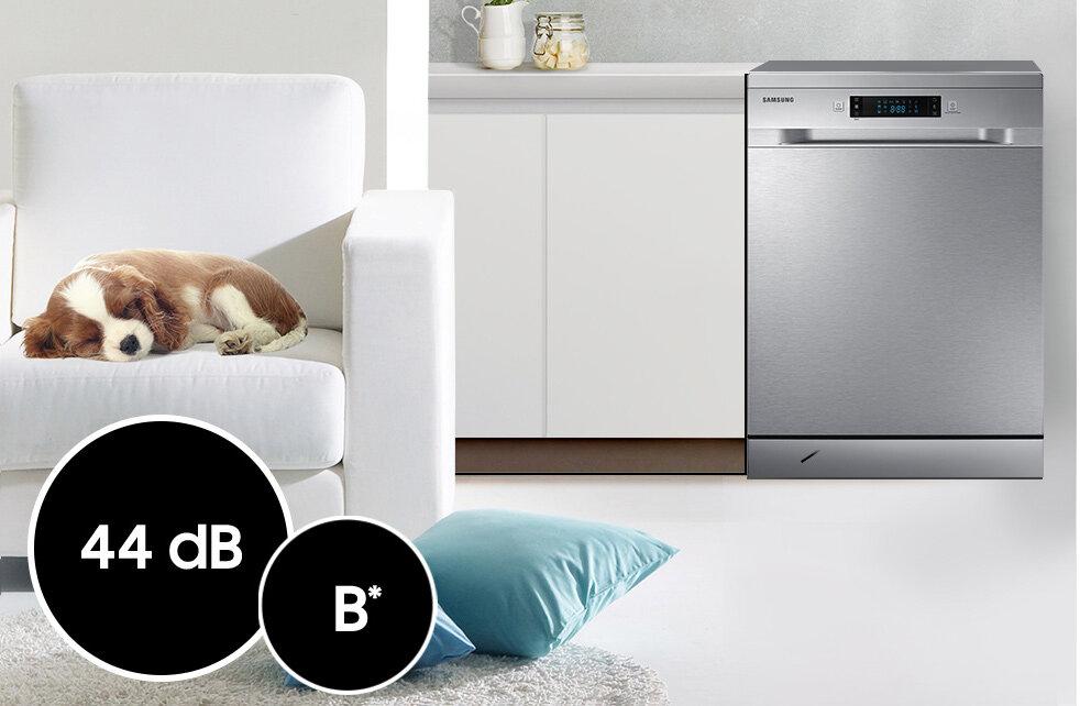 Na obrazie widoczna jest zmywarka w kolorze srebrnym z logo Samsung, umieszczona w kuchni obok białej szafki. Na pierwszym planie znajduje się biały fotel z psem oraz czarne koła z tekstem \'44 dB\' i \'B*\'.