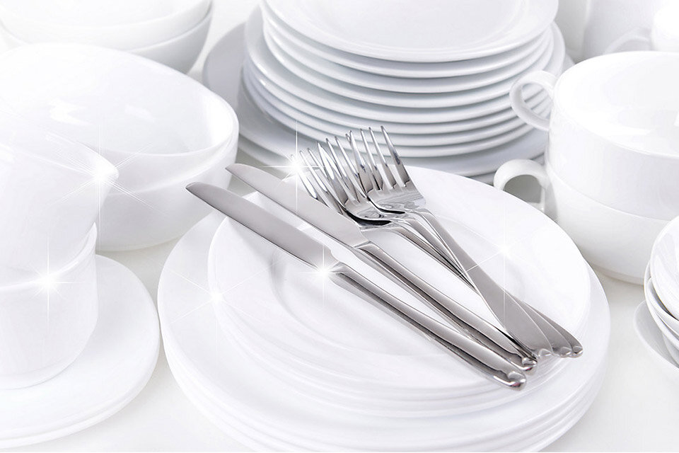 Na obrazie widoczne są białe talerze, filiżanki i miski ułożone w stosy, a na pierwszym planie leżą sztućce: noże i widelce. Naczynia i sztućce błyszczą, co sugeruje ich czystość.