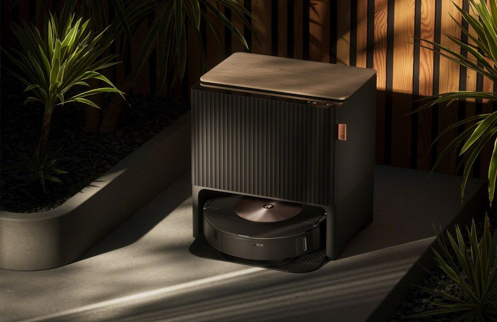 Na obrazie widoczny jest robot sprzątający w stacji dokującej, otoczony roślinami doniczkowymi. Stacja ma ciemny kolor z drewnianym elementem na górze.