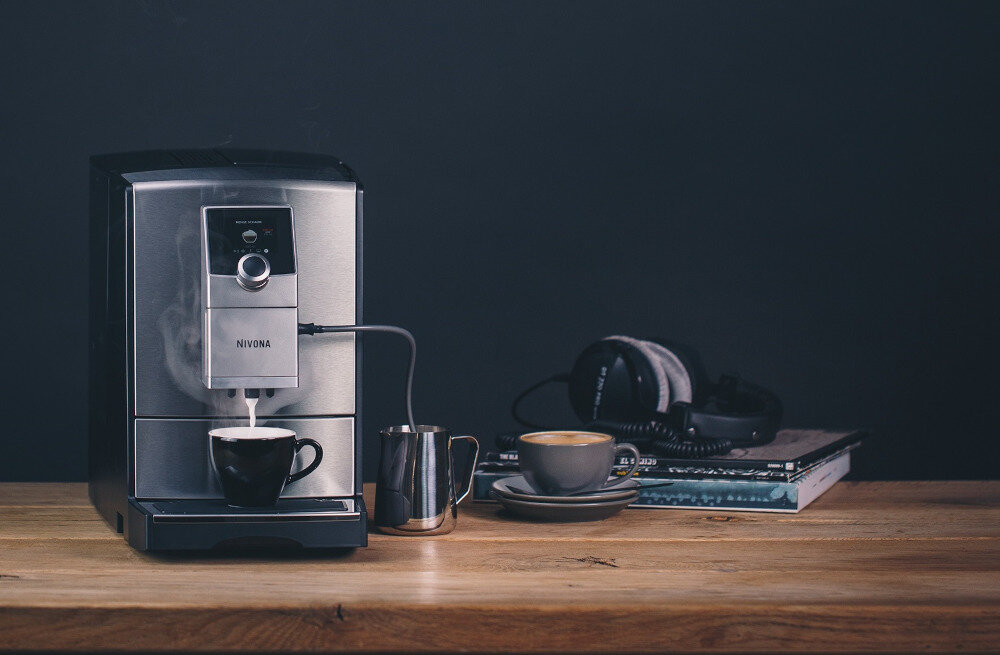 Na drewnianym stole stoi ekspres do kawy z logo NIVONA, obok filiżanka z kawą i metalowy dzbanek. W tle widoczne są słuchawki i stos książek.