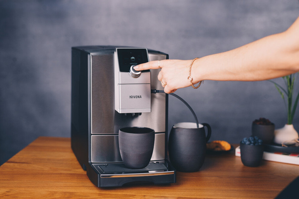 Ekspres do kawy z napisem \'NIVONA\' na froncie, z filiżanką ustawioną pod dyszą. Osoba naciska przycisk na panelu sterowania urządzenia.