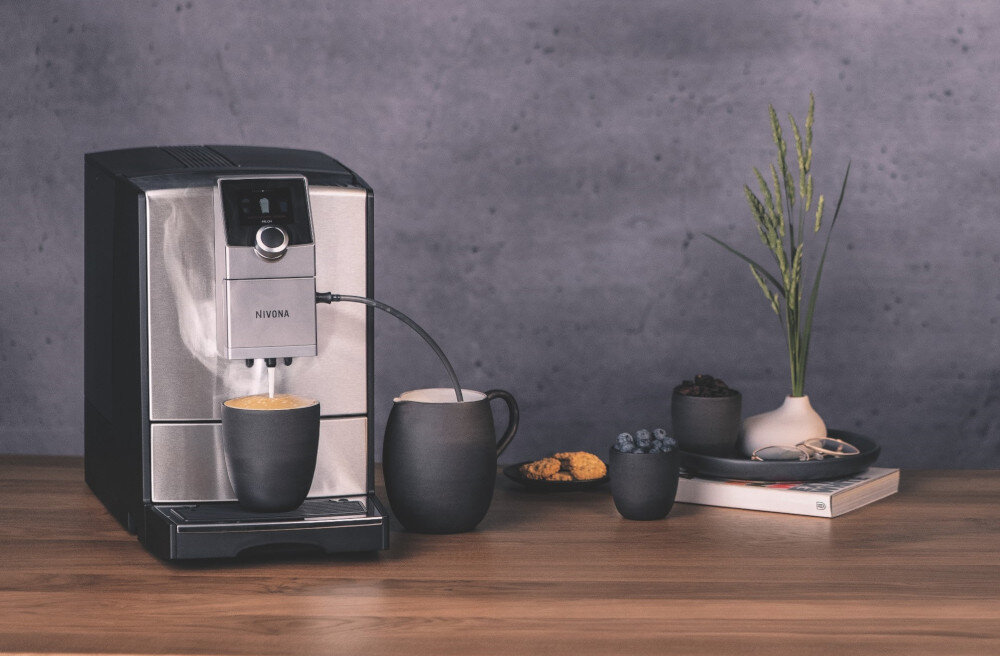 Na drewnianym blacie stoi ekspres do kawy z logo NIVONA, obok którego znajdują się filiżanki z kawą, talerzyk z ciastkami, miseczka z jagodami oraz wazon z zielonymi gałązkami.
