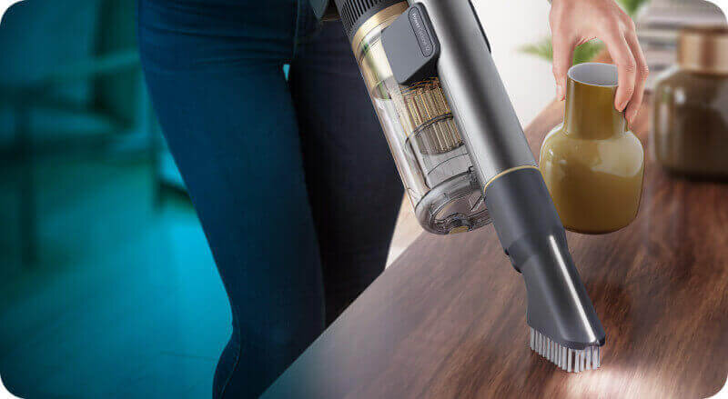 Osoba używa odkurzacza ręcznego na drewnianej powierzchni, obok stoi zielony wazon. Odkurzacz ma przezroczysty pojemnik na kurz i szczotkę na końcu ssawki.