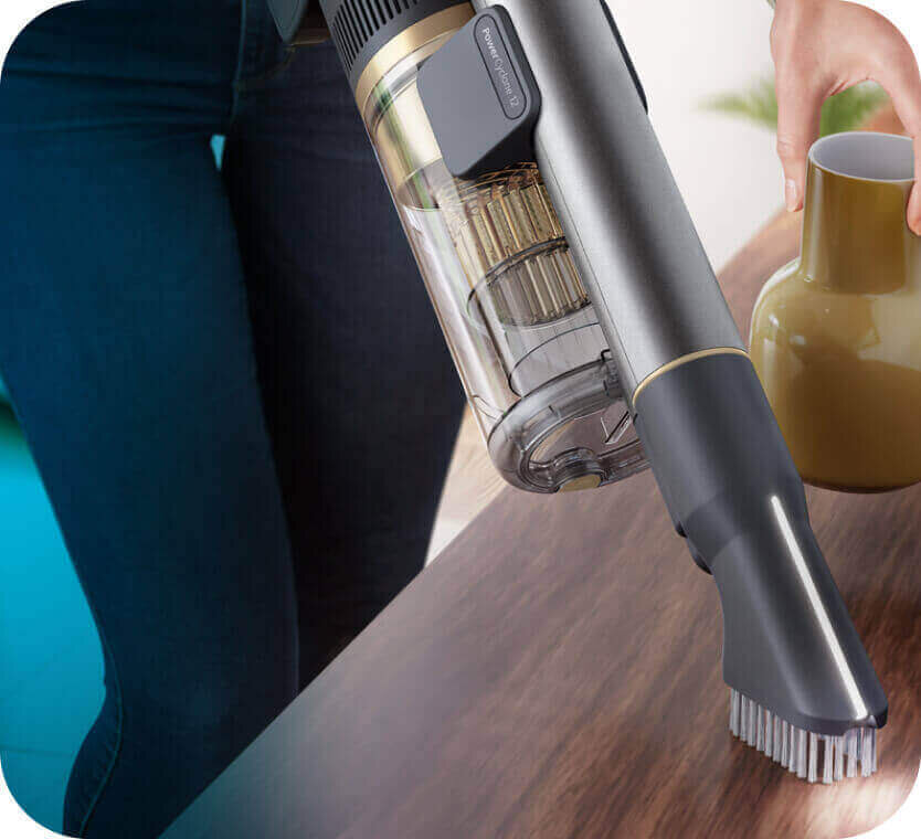 Osoba używa odkurzacza ręcznego z przezroczystym pojemnikiem na kurz do czyszczenia drewnianej powierzchni. Obok znajduje się ceramiczny wazon w kolorze brązowym.