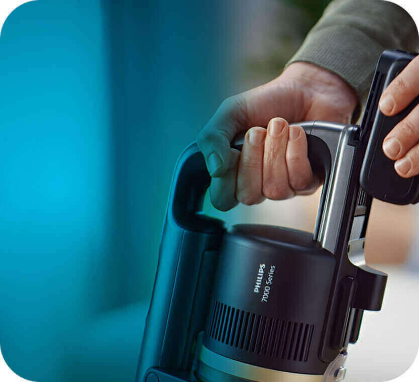 Osoba trzyma odkurzacz z widocznym napisem \'PHILIPS 7000 Series\' na obudowie. Tło jest rozmyte i ma niebieski odcień.
