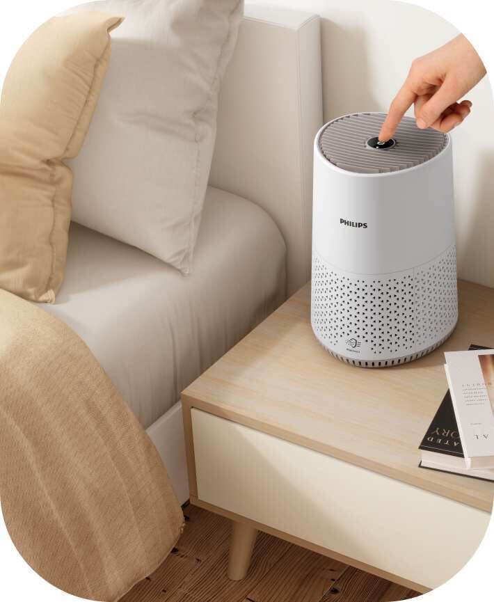 Na drewnianym stoliku nocnym obok łóżka stoi biały oczyszczacz powietrza z logo PHILIPS. Osoba naciska przycisk na górze urządzenia.