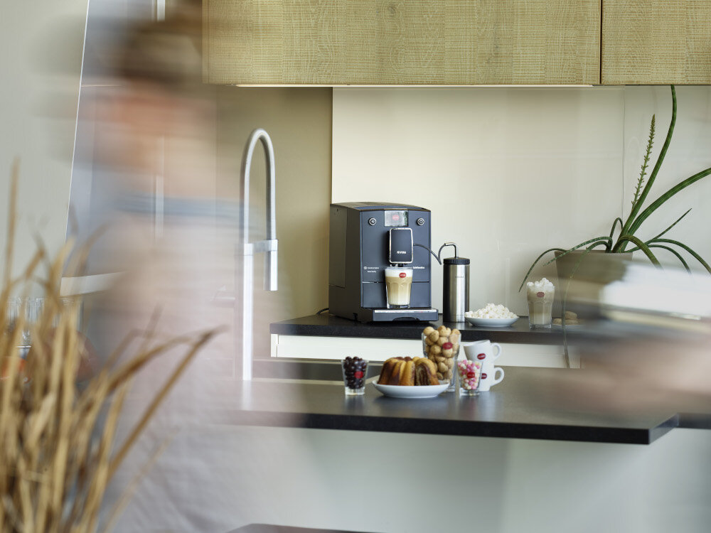 Na blacie kuchennym stoi ekspres do kawy z filiżanką pod wylewką, obok znajduje się pojemnik na mleko. Na blacie widoczne są również filiżanki, talerz z ciastkami i owoce, a w tle roślina doniczkowa.