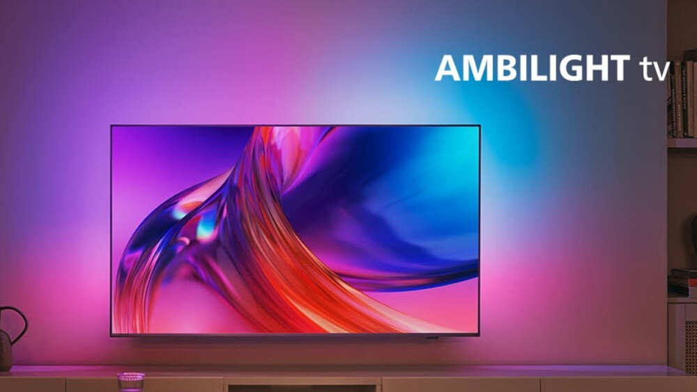 Telewizor wyświetla kolorowy obraz z efektem Ambilight, który rozświetla ścianę za nim. Na ekranie widoczny jest dynamiczny wzór w odcieniach fioletu i czerwieni, a w rogu znajduje się napis \'AMBILIGHT tv\'.