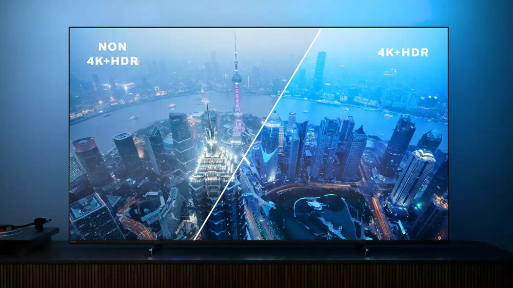 Telewizor wyświetla obraz miasta z widocznym podziałem na \'NON 4K+HDR\' i \'4K+HDR\'. W tle widać wieżowce i rzekę, a ekran emituje niebieskie światło.
