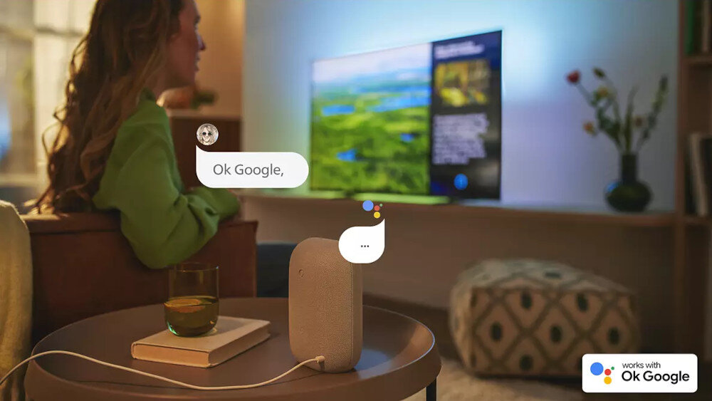 Osoba siedzi na kanapie przed telewizorem, na którym wyświetlany jest krajobraz. Na stoliku obok znajduje się inteligentny głośnik, a w rogu ekranu widnieje logo \'works with Ok Google\'.