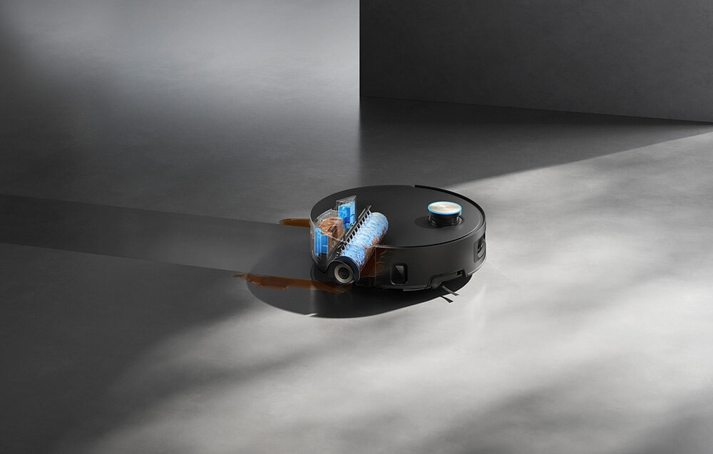 Czarny robot sprzątający na szarej podłodze, z widocznym przezroczystym elementem pokazującym wewnętrzny mechanizm z niebieskim wałkiem. Robot znajduje się w oświetlonym miejscu, a reszta podłogi jest w cieniu.