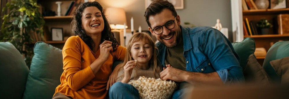 Trzy osoby siedzą na kanapie, uśmiechają się i jedzą popcorn. W tle widać elementy wystroju wnętrza, takie jak rośliny i półki z książkami.