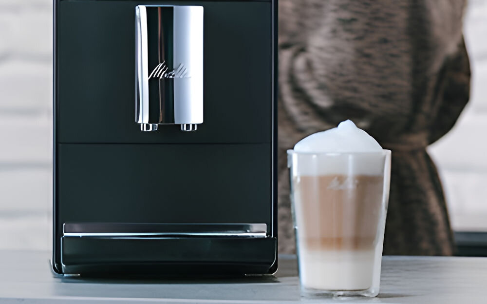Czarny ekspres do kawy z logo \'Melitta\' na metalowej części. Obok stoi szklanka z warstwową kawą i pianką na wierzchu.