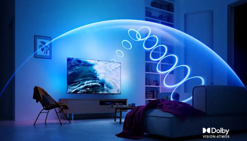 W pokoju znajduje się telewizor wyświetlający obraz fal morskich, otoczony niebieskim światłem. W prawym dolnym rogu widnieje logo \'Dolby Vision-Atmos\'.