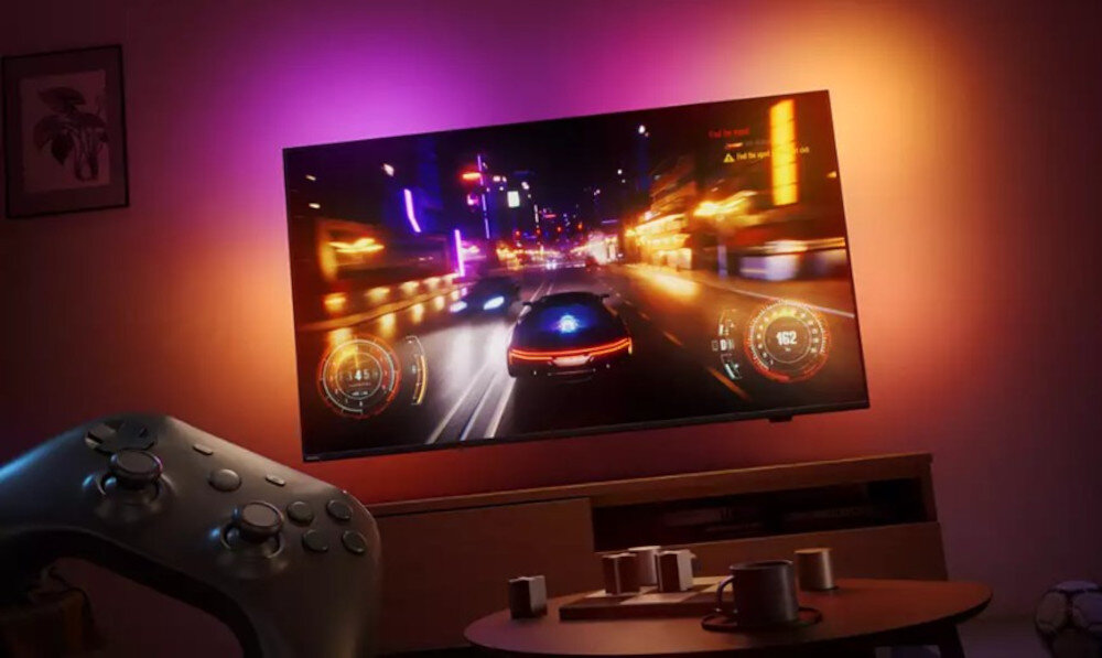 Telewizor wyświetla dynamiczną scenę z gry wyścigowej, otoczony kolorowym oświetleniem. Na stoliku przed telewizorem znajdują się filiżanki, a w pobliżu widoczny jest kontroler do gier.