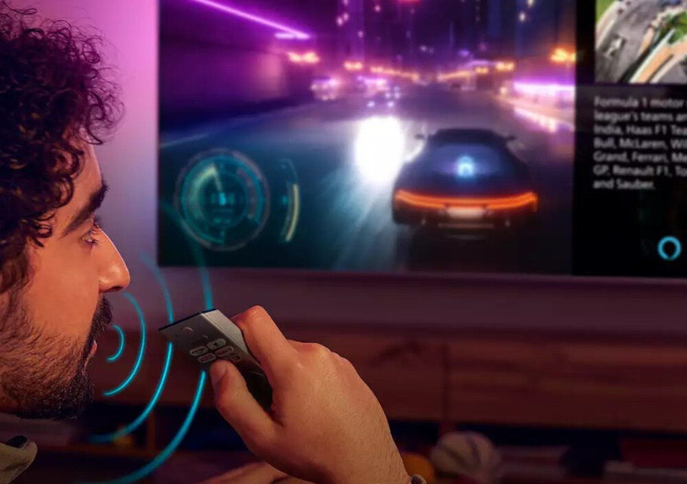 Osoba trzyma pilot przed telewizorem, na którym wyświetlana jest dynamiczna scena z gry wyścigowej. Obok widoczny jest tekst dotyczący zespołów Formuły 1, w tym India, Haas F1 Team, Ferrari, McLaren, Williams, Mercedes GP, Renault F1 Team i Sauber.