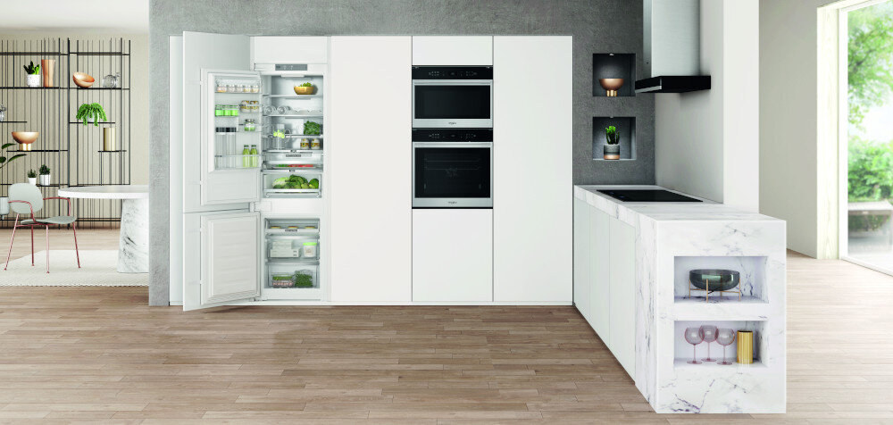 Kuchnia z białymi szafkami, w której znajduje się otwarta lodówka z widocznymi produktami spożywczymi. Obok lodówki znajdują się dwa piekarniki w zabudowie, a na blacie widoczne są dekoracyjne elementy.