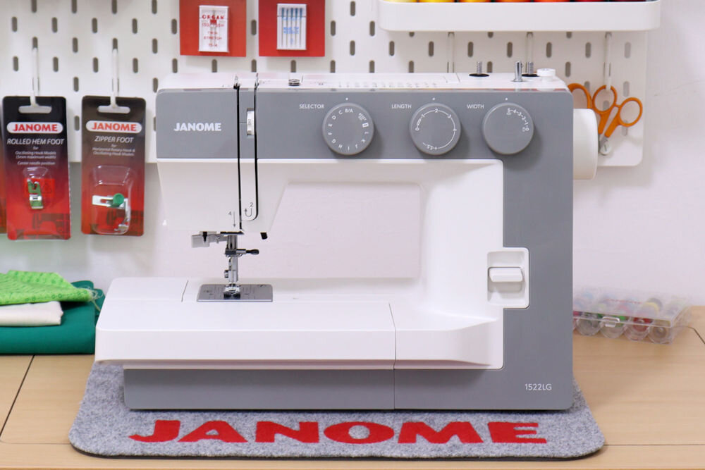 Maszyna do szycia JANOME 1522LG w kolorze szaro-białym z trzema pokrętłami do regulacji. W tle widoczne są akcesoria do szycia oraz napis \'JANOME\' na macie.