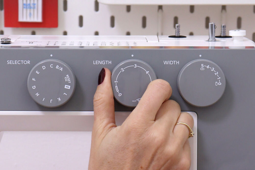 Zdjęcie przedstawia panel maszyny do szycia z trzema pokrętłami oznaczonymi jako SELECTOR, LENGTH i WIDTH. Dłoń reguluje pokrętło LENGTH, a w tle widoczne są akcesoria do szycia.