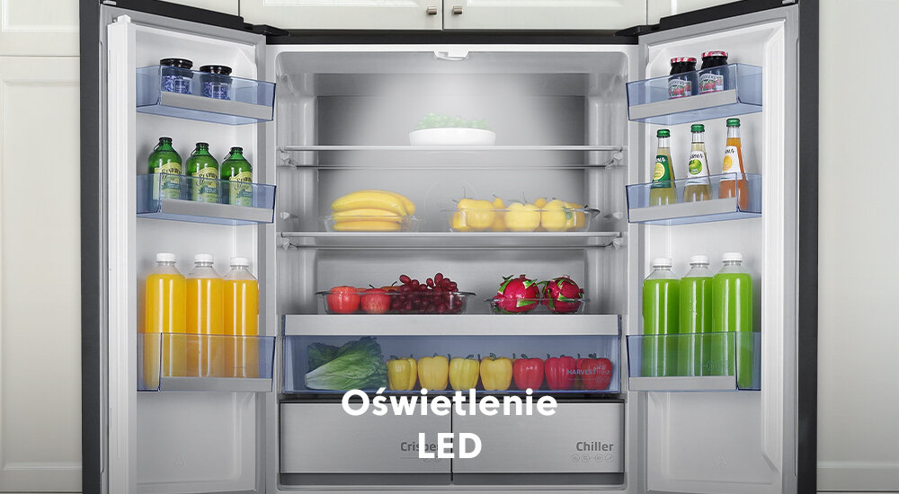 Otwarta lodówka z widocznymi półkami wypełnionymi różnymi produktami spożywczymi, takimi jak napoje, owoce i warzywa. Na obrazie widoczny jest tekst: \'Oświetlenie LED\'.
