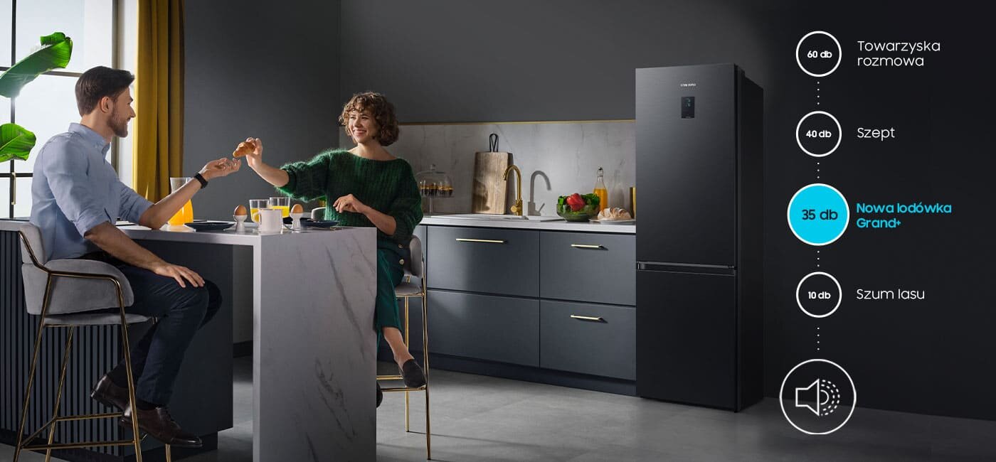 W kuchni znajduje się czarna lodówka SAMSUNG z napisem \'Nowa lodówka Grand+ 35 dB\'. Obok lodówki siedzi para przy stole, pijąc napoje.