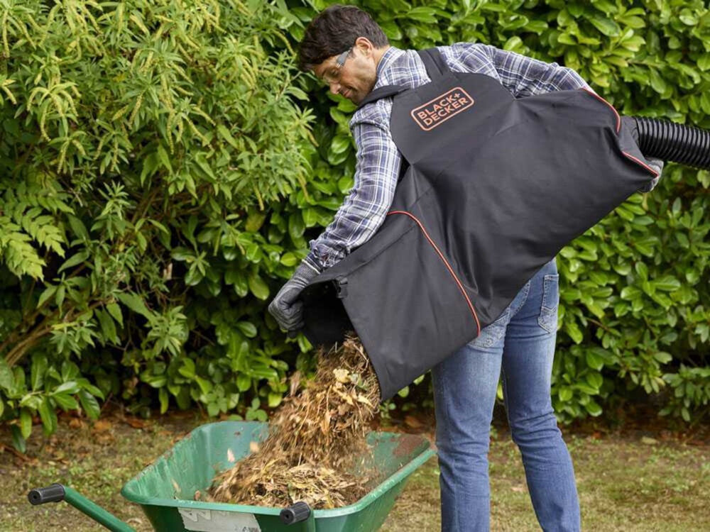 Osoba opróżnia worek z liśćmi do taczki, nosząc torbę z logo \'BLACK+DECKER\'. W tle widoczne są zielone krzewy.
