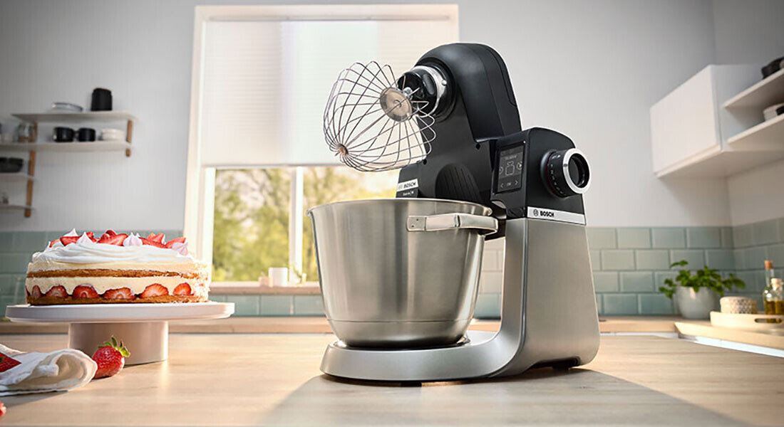 Na blacie kuchennym stoi robot kuchenny z metalową misą i trzepaczką. W tle widać tort z truskawkami oraz okno z widokiem na zewnątrz.