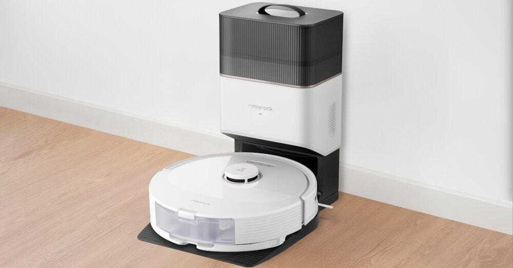Na podłodze znajduje się biały robot sprzątający z okrągłą obudową, stojący na stacji dokującej. Na stacji widoczne jest logo \'roborock\'.