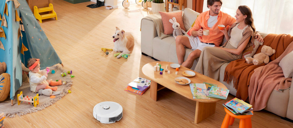 Na drewnianej podłodze w salonie znajduje się biały robot sprzątający. W tle widać rodzinę siedzącą na kanapie oraz bawiące się dziecko i psa.