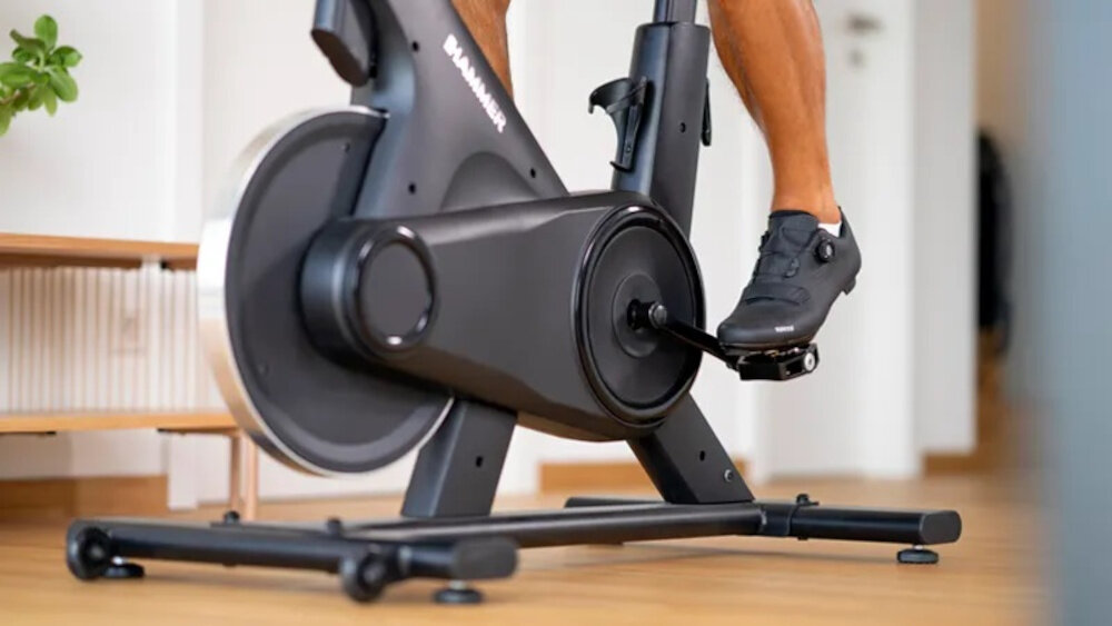 Osoba korzysta z roweru spiningowego w pomieszczeniu. Na ramie roweru widoczne jest logo \'HAMMER\'.