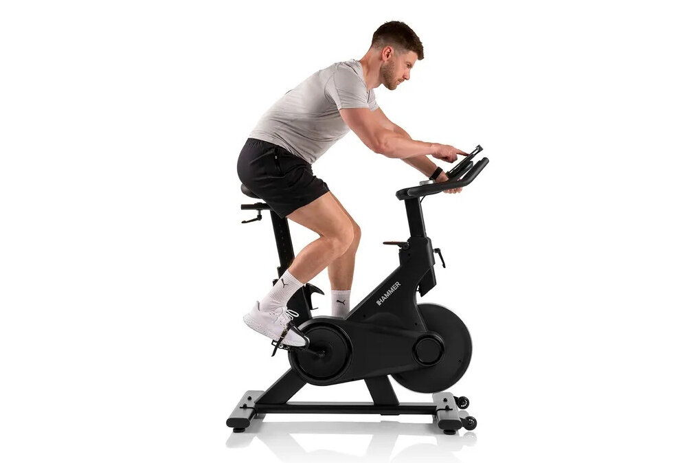 Osoba ćwiczy na rowerze spinningowym HAMMER Speed Race X. Rower ma czarną ramę i wyświetlacz na kierownicy.
