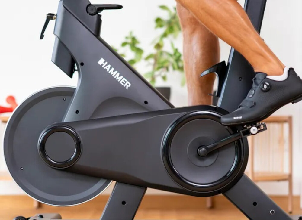 Osoba ćwiczy na rowerze spiningowym marki HAMMER, widoczny jest mechanizm napędowy i pedały. W tle znajduje się roślina doniczkowa i mebel.